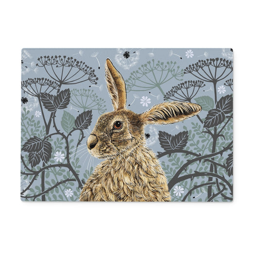Our rectangular blue hare tempered glass chopping board features a hare portrait with one floppy ear, around the hare are stylised brambles, leaves, cow parsley and dandelion puffs, background colour is a blue  grey.